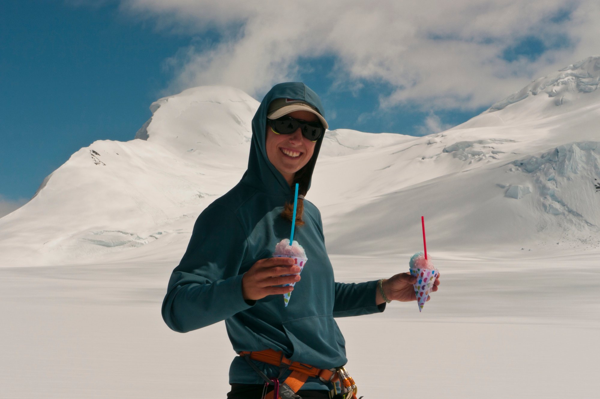 smiling NOLS participant holds a snow cone in each hand with snowy mountains in the background