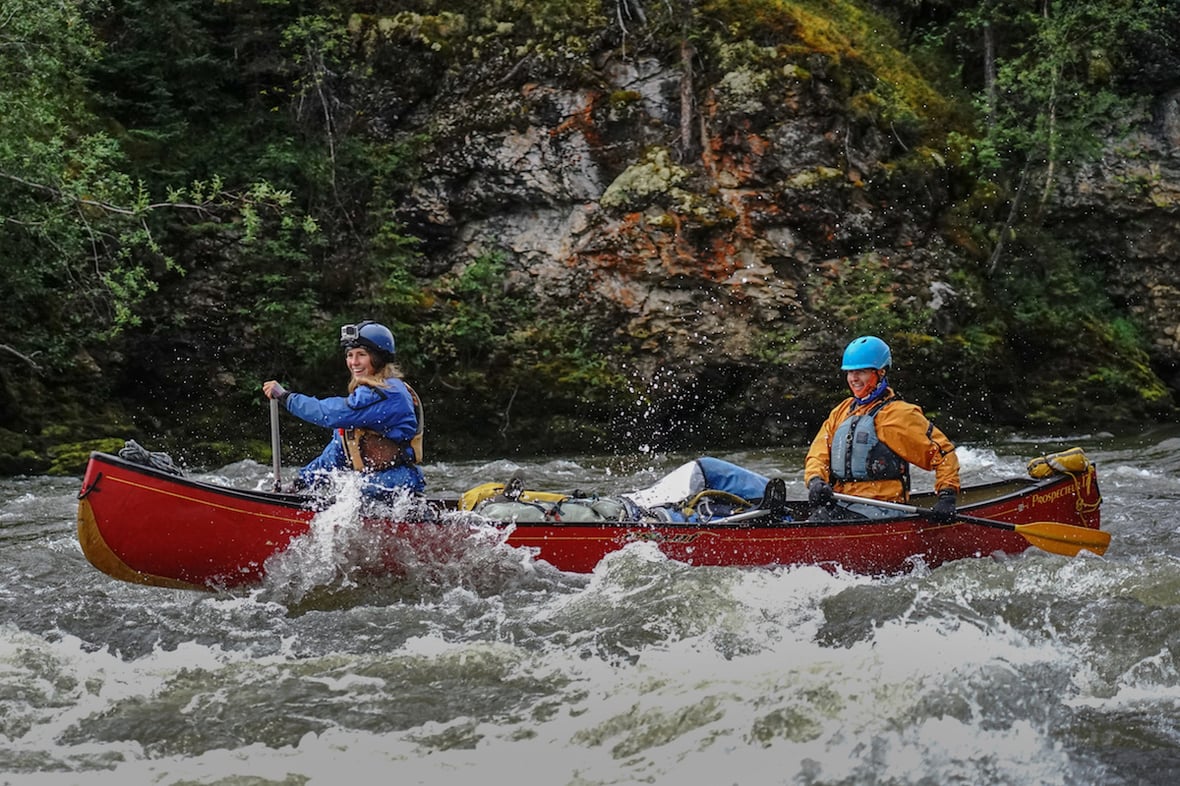 two smiling students wearing helmets paddle a red canoe through a rapid in the Yukon