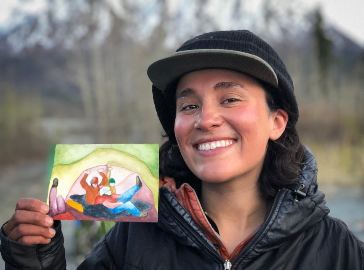 smiling female NOLS participant wearing black jacket and hat over baseball cap holds up a brightly colored watercolor painting in the mountains