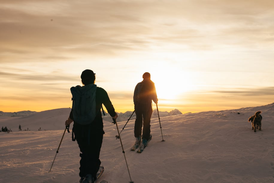 two backcountry skiers make their way to the top of a slope at sunset, accompanied by a small dog