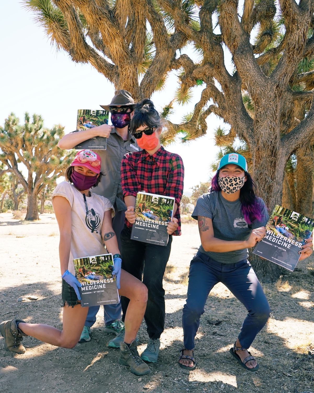 Four NOLS students smile while wearing masks and hold up the Wilderness Medicine Handbook