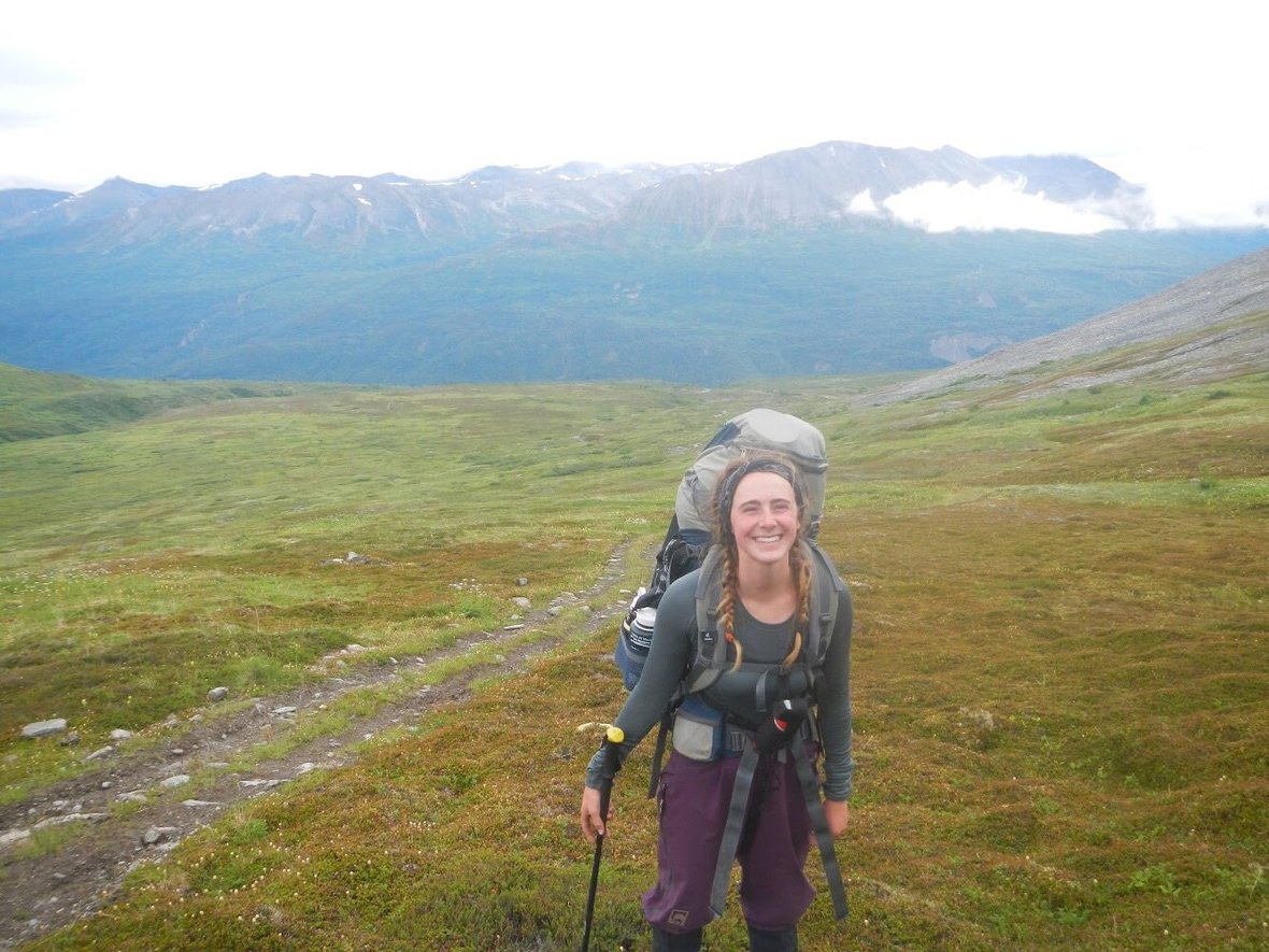 smiling woman with backpack and braided hair pauses in a green valley while backpacking in Alaska