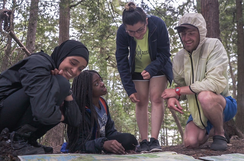 four NOLS participants sit and bend over to look at a map spread out on the ground in a forest in the Adirondacks