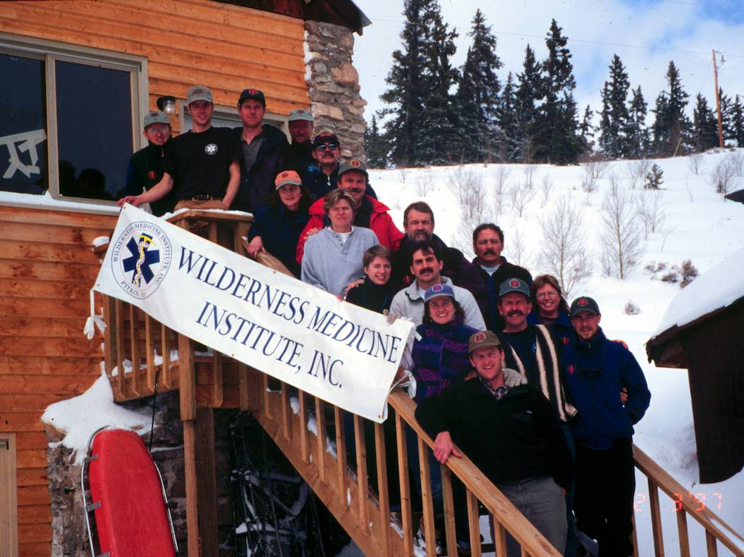Group picture of staff in front of the early WMI Staff Building holding a banner that says "Wilderness Medicine Institute, Inc."