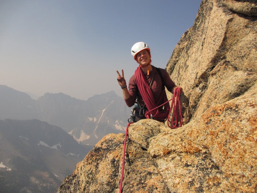 Climber smiles and shows a peace sign while coiling rope partway up an alpine climb