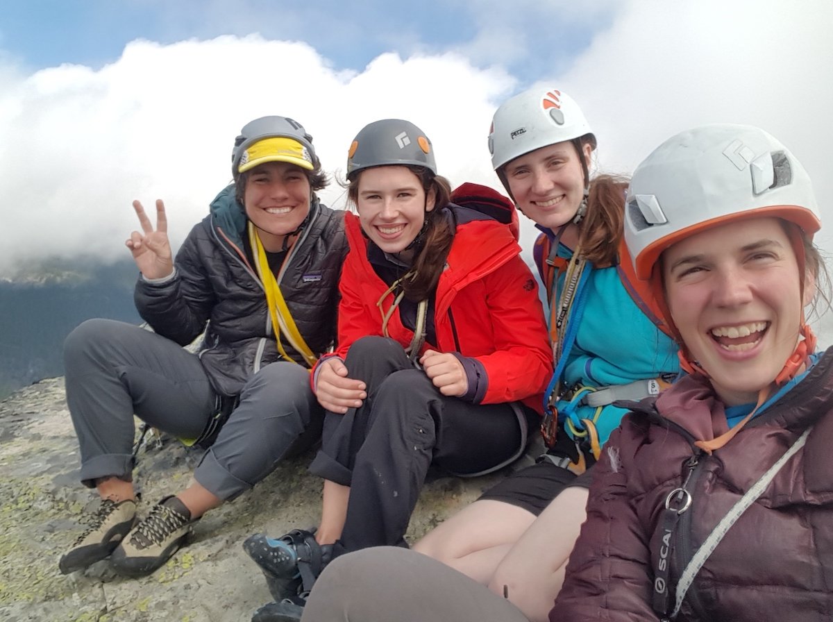 Four climbers smile wearing climbing helmets on an alpine rock summit