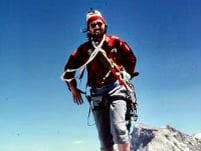 Peter Simer in red and black checkered shirt and stocking cap with a climbing rope across his shoulders as he summits a peak.