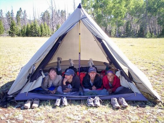 four smiling teenage girls lie on their tummies looking out of a tan tent in the mountains