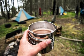 hand holding cup of coffee with NOLS tents in background