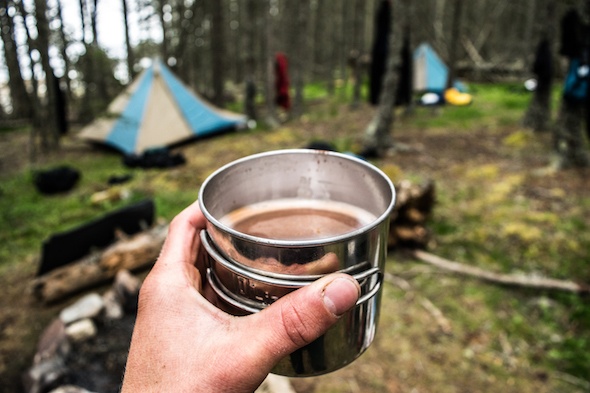 hand holding a cup of coffee with NOLS tents amongst the trees in the background