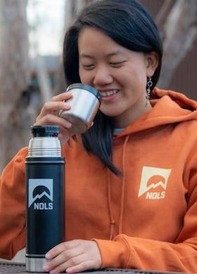 smiling person in NOLS sweatshirt drinks from NOLS thermos