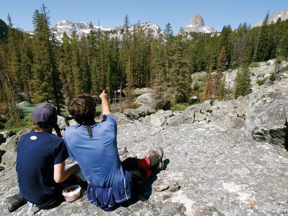 two NOLS students sit on rocky outcrop and look at a view of trees and snow-capped mountains in the Rockies