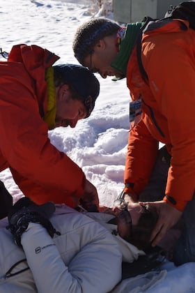 two men in orange jacket care for a patient lying in the snow on a WFR course