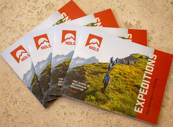 four NOLS expeditions catalogs with orange logo and people hiking on the cover fanned out on a tan floor