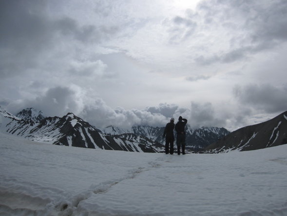 two silhouetted figures stand on a snowfield in Alaska and look out toward mountains shrouded in clouds