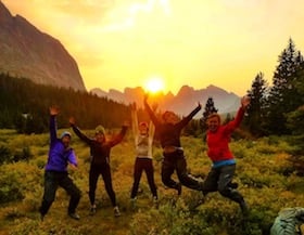 five smiling NOLS students jump into the air in the mountains with sun setting behind