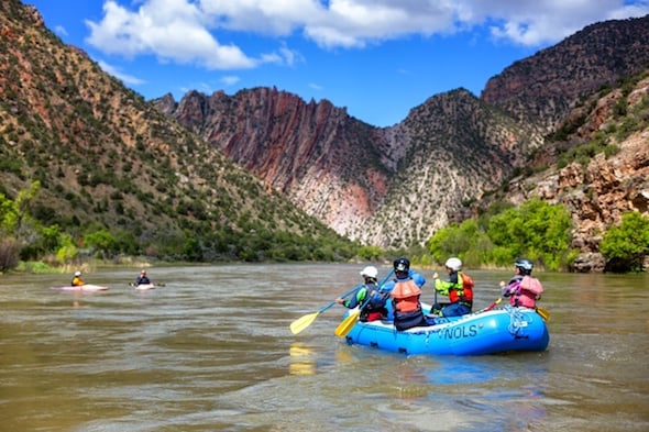 four NOLS students paddle a raft on a quiet section of river with redrock canyon walls beside