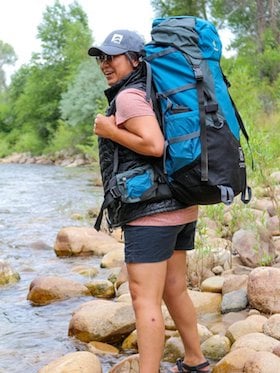 smiling NOLS participant stands at edge of stream wearing NOLS hat and backpack