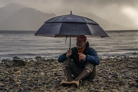 NOLS instructor sits on rocky beach in Patagonia holding umbrella