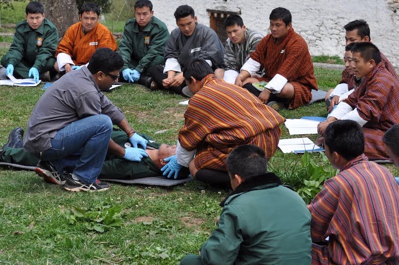 11 seated students look on as two wilderness medicine participants practice carrying for a patient lying on a mat in the grass