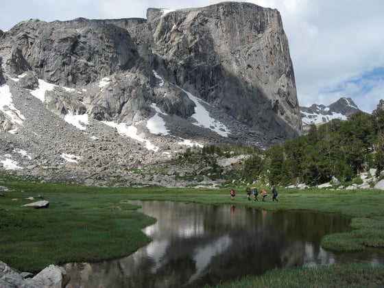 backpackers walk alongside a glassy alpine lake toward a rocky peak with permanent snowfields in the Wind River Range