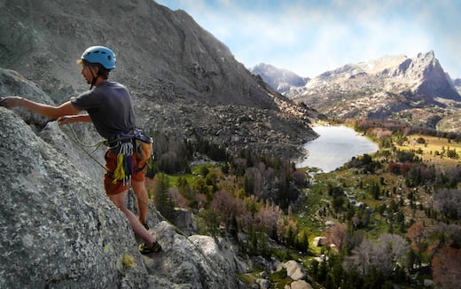 rock climber wearing blue helmet pauses on a rock face in the Rockies with alpine lake and more mountains beyond