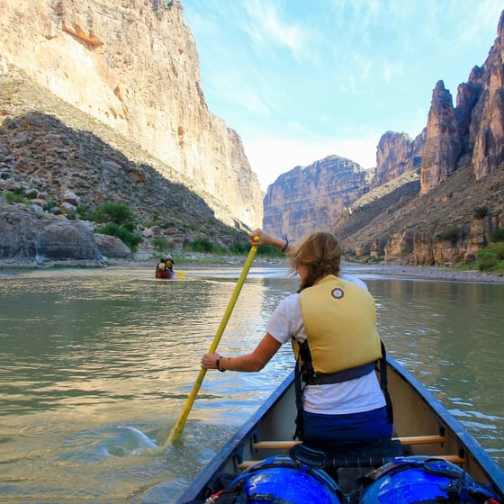 woman wearing yellow life preserver sits in the bow of a canoe while paddling in a river canyon
