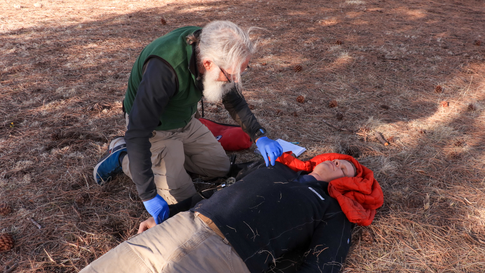 An older man checks the pulse of the an unconscious patient. The patient's neck is stabilized by a coat.