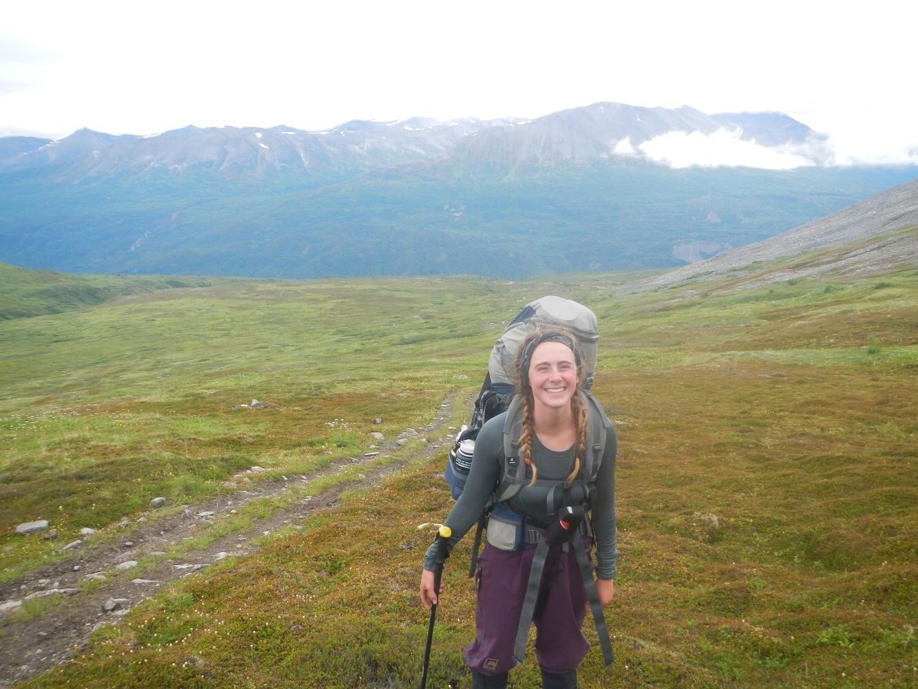 Woman with hair in braids wearing a backpack and smiling next to a trail in Alaska's mountains
