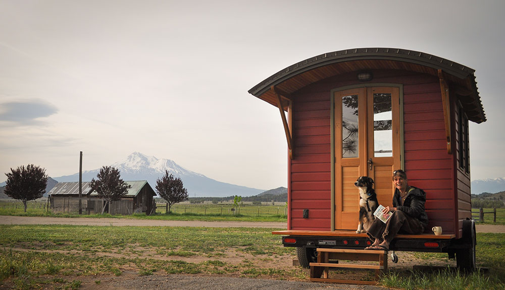smiling woman with dog sitting on the porch of a red tiny house with mountains in the background