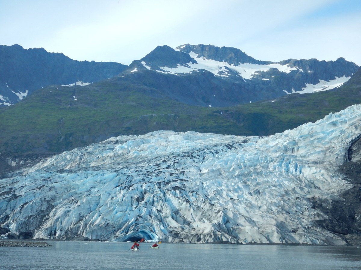 Two sea kayakers paddle on the coast with large glacier and green coastline in the background