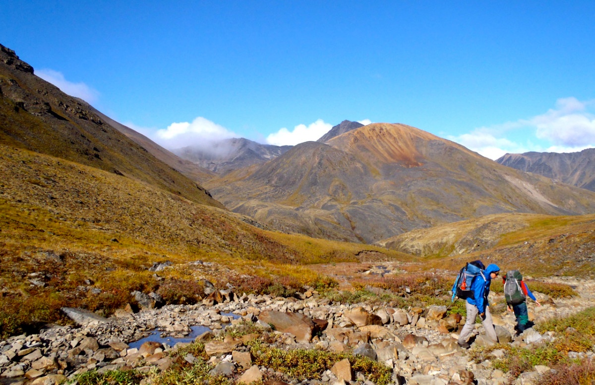 two NOLS students hike across rocky terrain near a creek in the mountains of Alaska