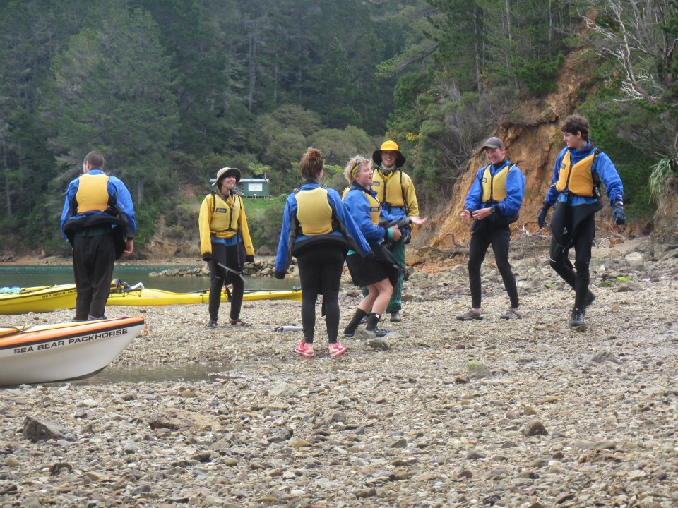 Smiling NOLS students wearing yellow life preservers dance on a pebbly beach in New Zealand