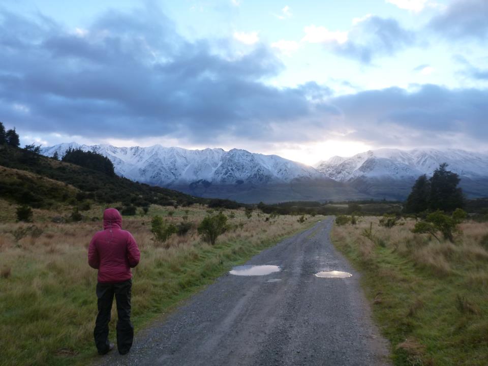 Student in a pink jacket looks down a gravel path heading toward snowy peaks as the sun sets behind clouds