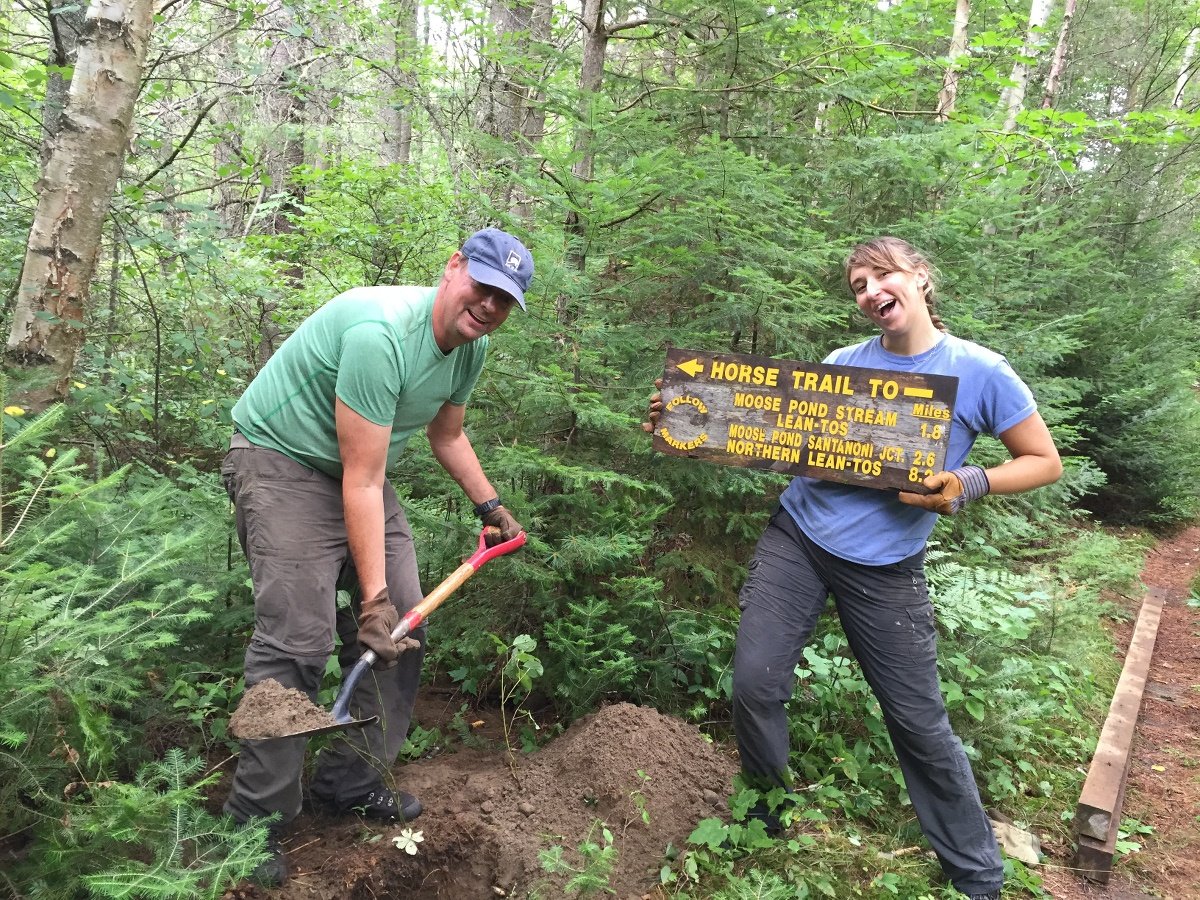 two smiling people work on a service project with man using a shovel and woman holding a trail sign