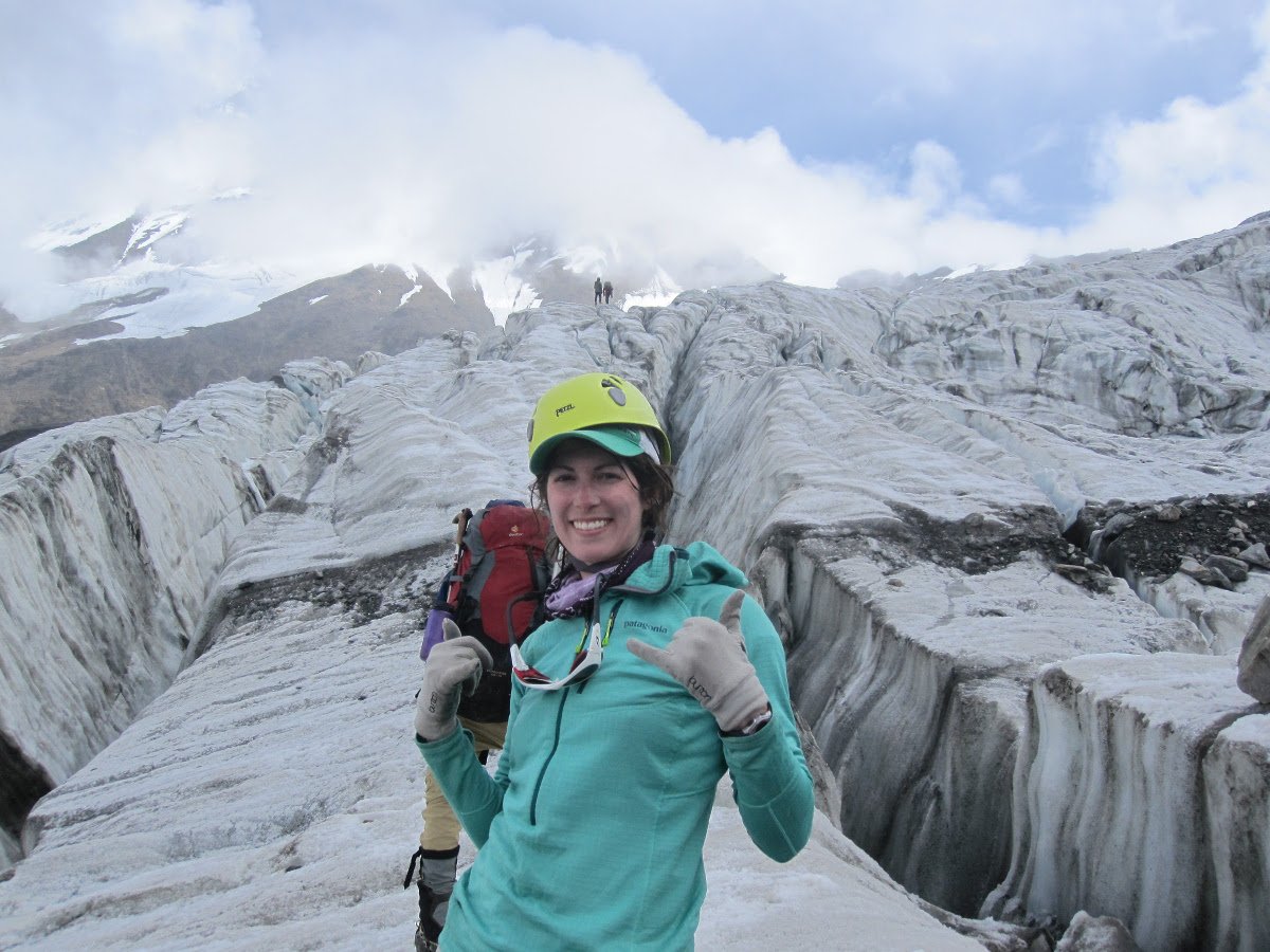 Smiling NOLS student wearing helmet and hat pauses on a glacier for a photo