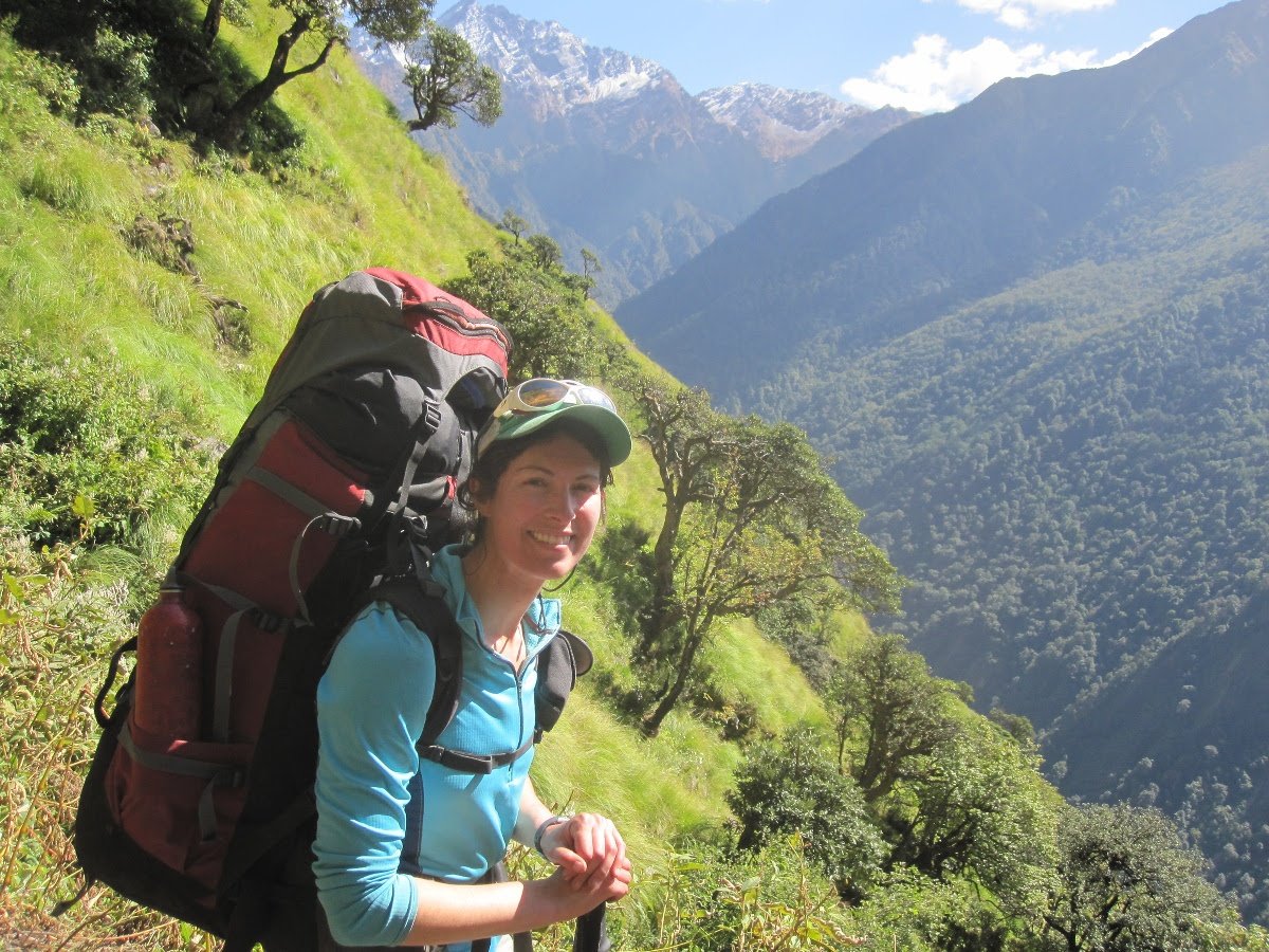 Smiling woman wearing a red backpack leans on a trekking pole while walking on a steep green slope in the mountains
