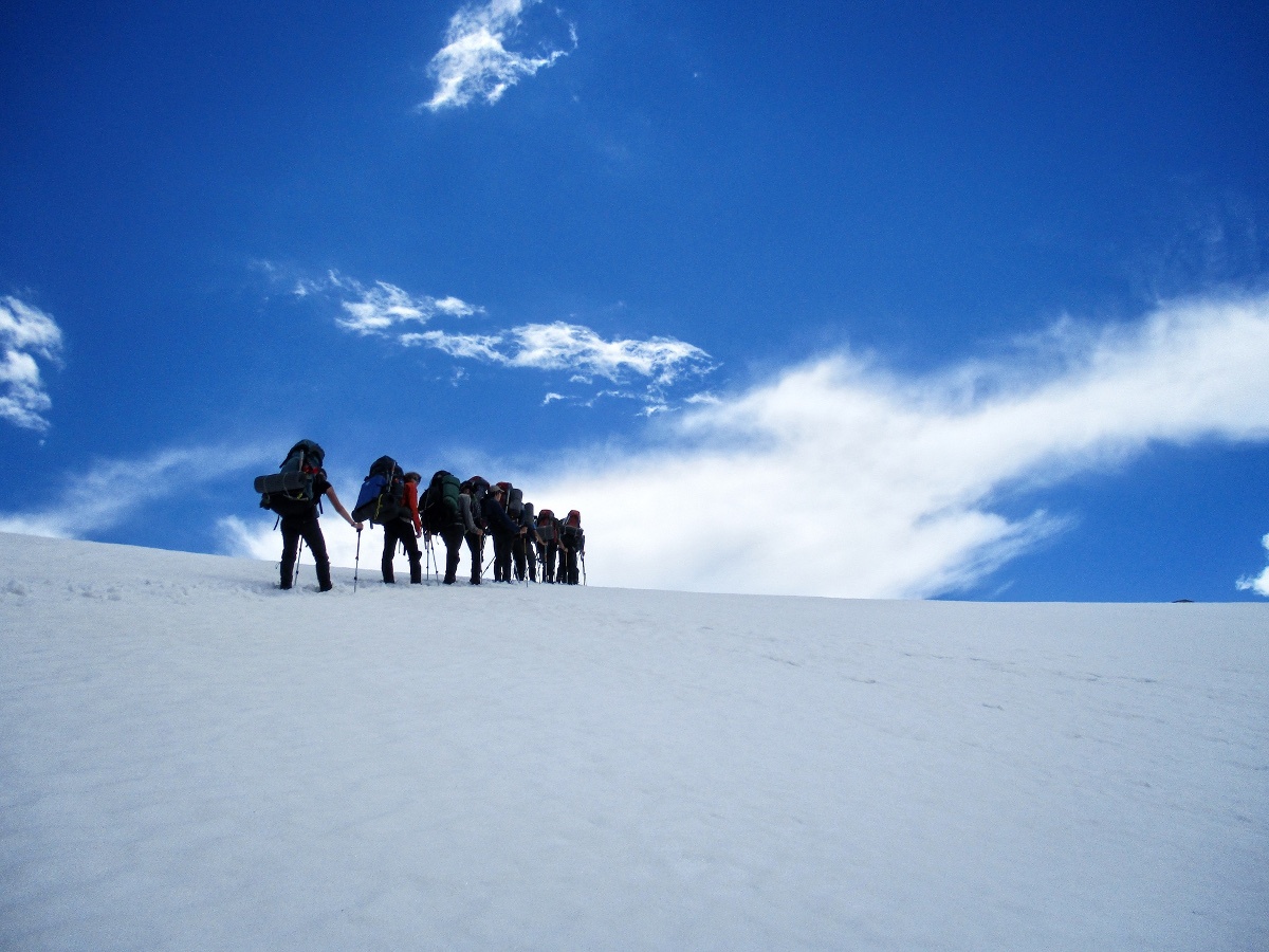 line of hikers wearing backpacks trek up a snowfield with blue sky and clouds above
