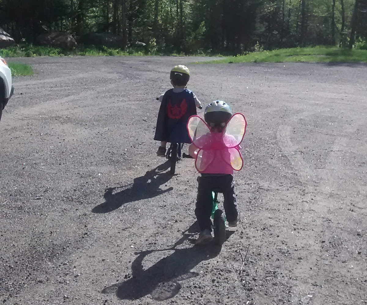 two small children riding bikes, one with superhero cape and one with pink butterfly wings, both wearing helmets