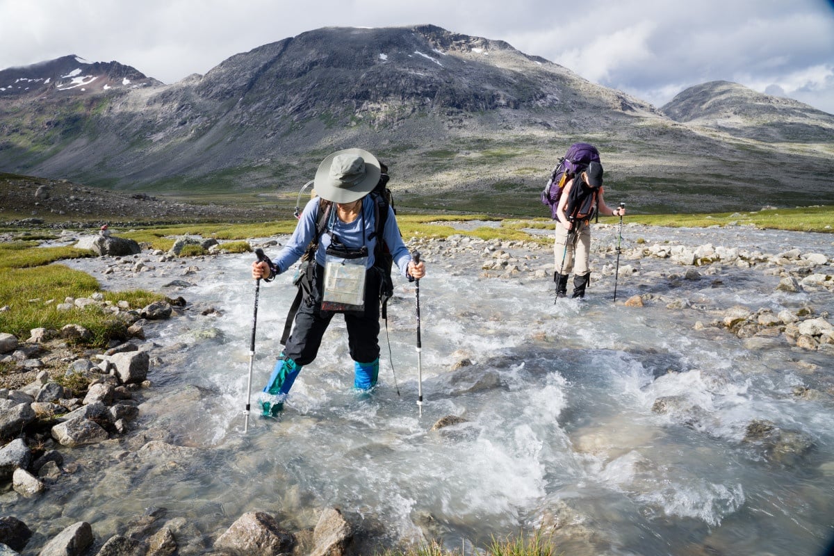 Two people cross a river using trekking poles