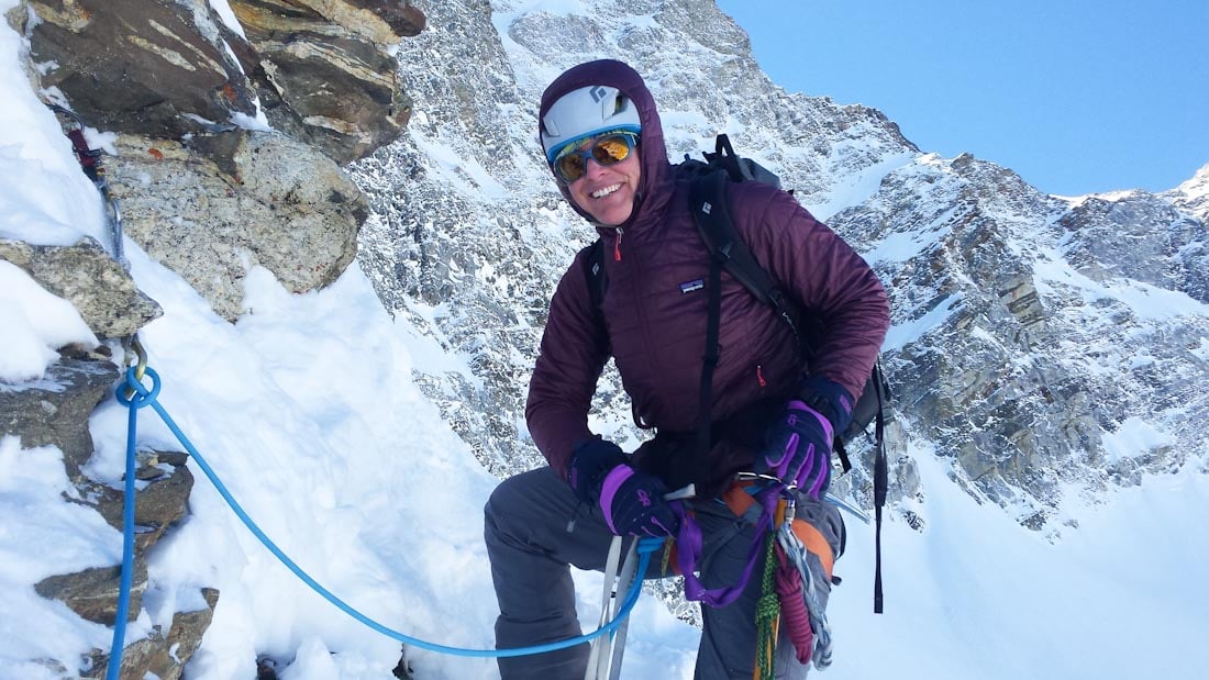 Jaime Musnicki poses wearing a jacket and climbing harness and tied into climbing protection on a snowy route