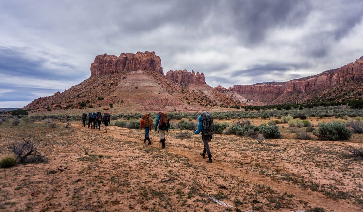 NOLS students hike single file along reddish dirt toward red rock formations in Escalante
