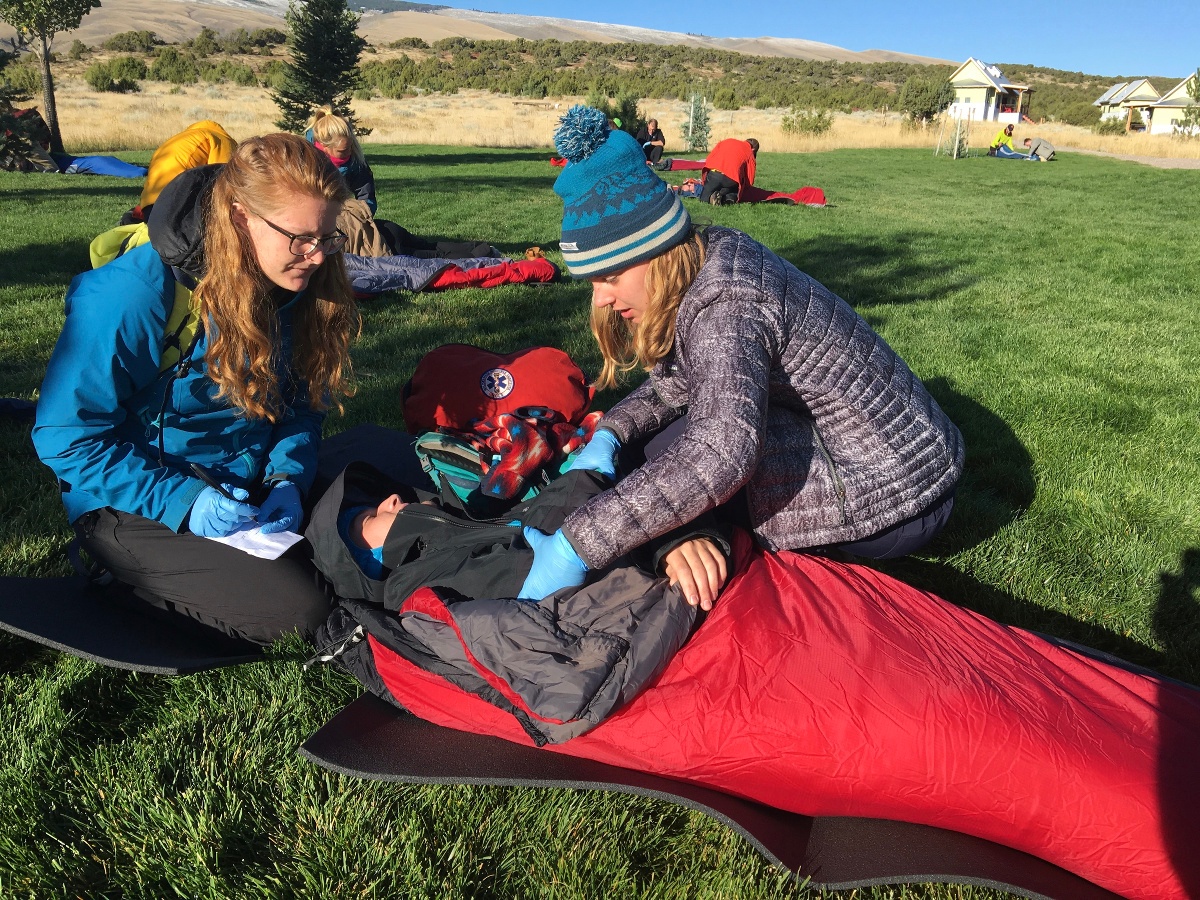 two female wilderness medicine students practice caring for a patient lying in a sleeping sleeping bag outdoors