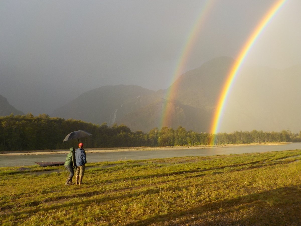 Two people with an umbrella watch two rainbows in the distance arc over a river