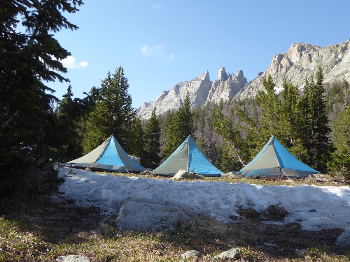 Three NOLS tents set up in a grassy clearing surrounded by trees and mountains