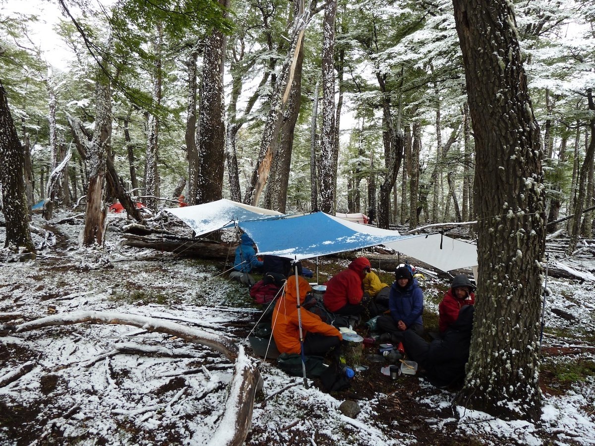 Students huddle under tarps to cook in a forest dusted with snow