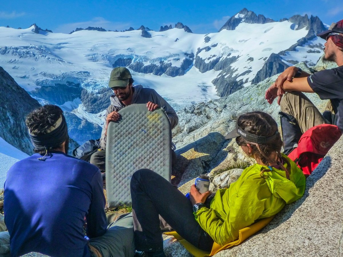 NOLS instructor holding foam pad teaches a lesson to students seated on a rocky outcrop in snowy mountains