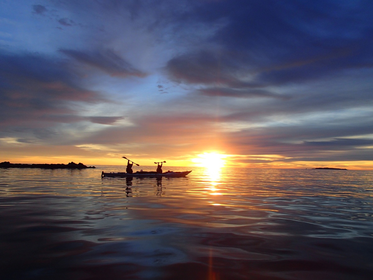 Two people in a tandem kayak raise paddles with the sunrise behind them while floating on the ocean