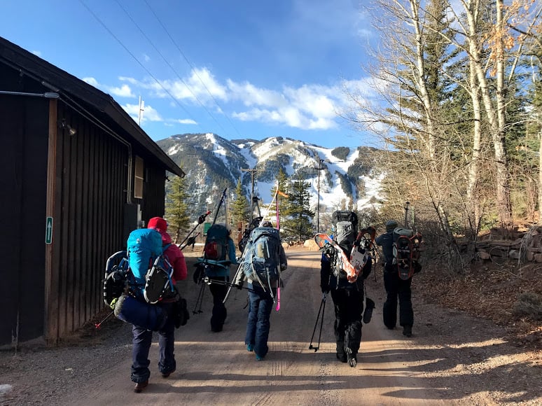 A group walks towards mountains on a dirt road carrying backcountry ski and snowshoe equipment