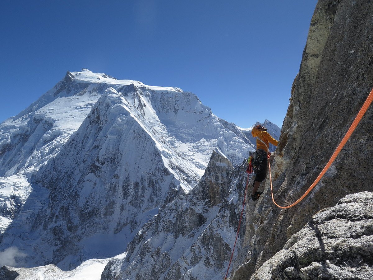 rock climber in a harness on a steep face surrounded by snowy high-altitude peaks